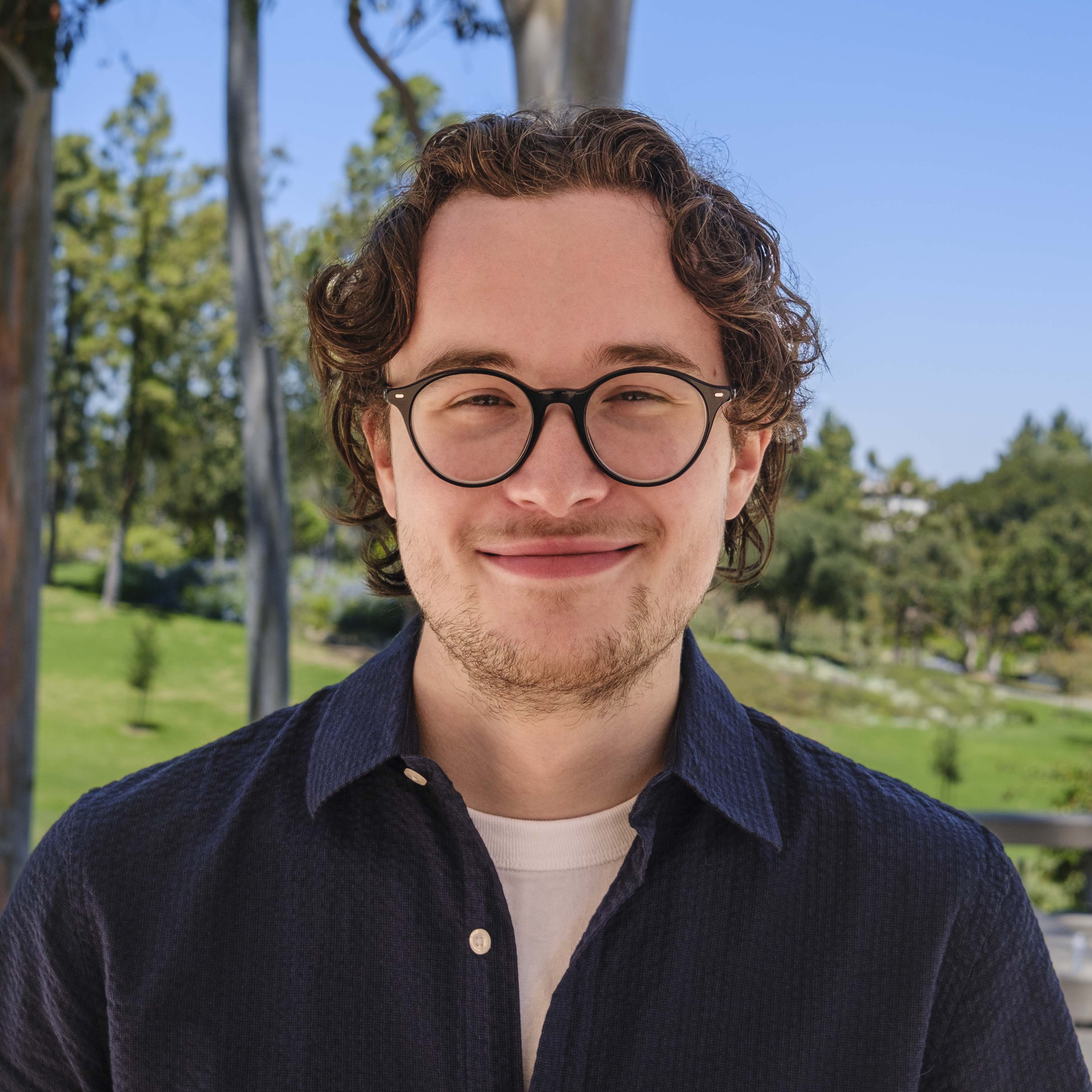 Young man with curly brown hair and round glasses smiles at the camera, wearing a navy button-up shirt over a light t-shirt in an outdoor setting with trees and blue sky in the background.